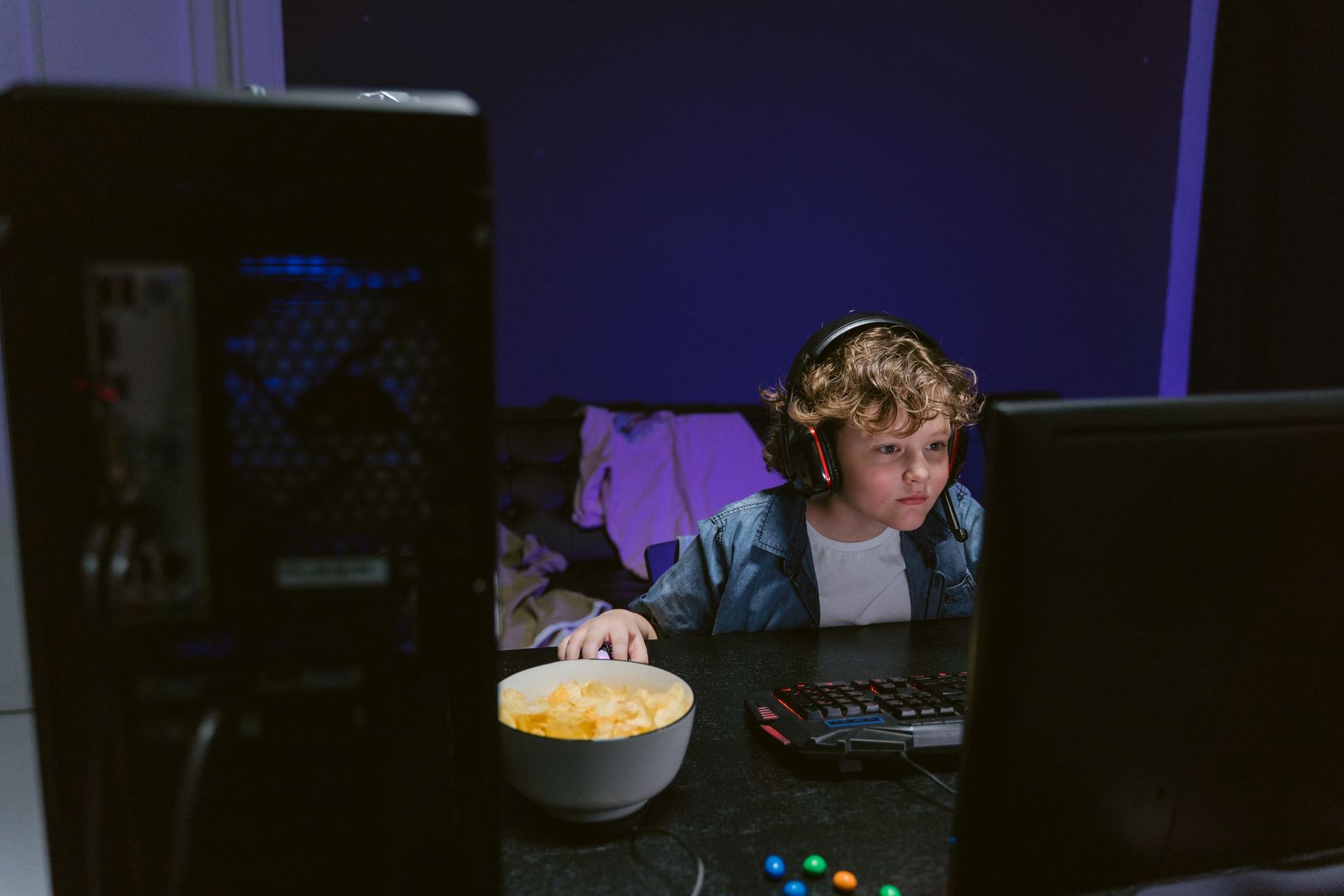 A boy wearing a headset sits at a computer in a dim room