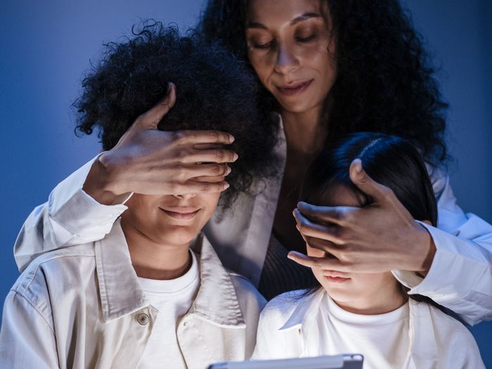 A mother gently covers her children’s eyes while they look at a tablet